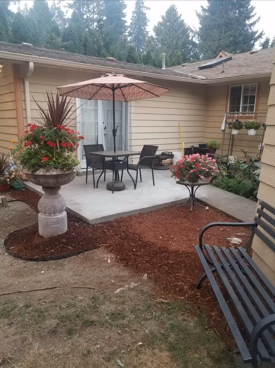 Outdoor patio area with a concrete slab featuring a table, four chairs, and a large umbrella. There are two large flower pots with colorful flowers and plants, a black metal bench, and a garden area with mulch. The patio is adjacent to a beige building with windows and surrounded by trees in the background.