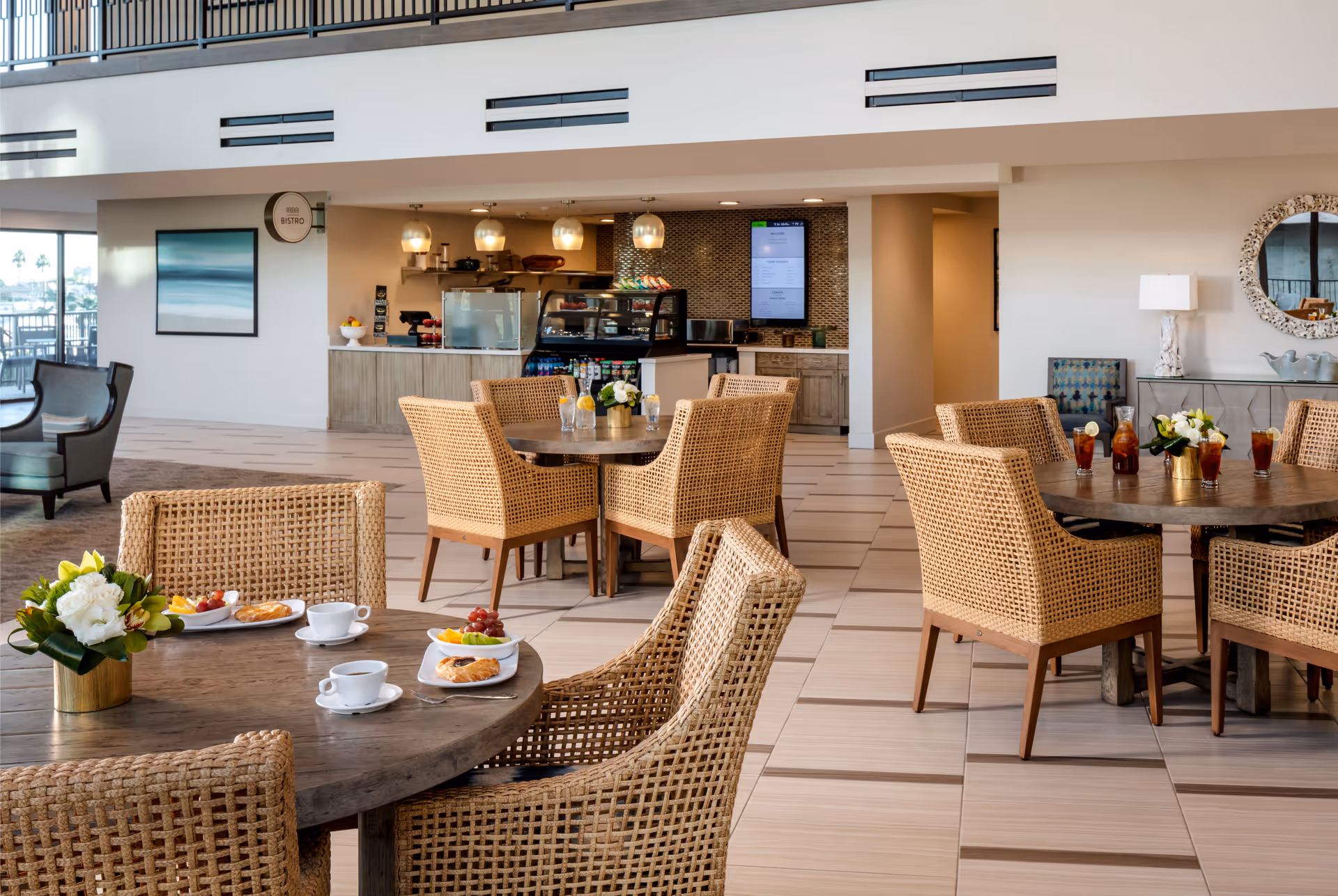 A bright and spacious dining area in a senior living facility with round wooden tables surrounded by wicker chairs. The tables are set with plates of food, cups, and glasses of iced tea. In the background, there is a small bistro counter with hanging pendant lights, a digital menu screen, and a display case with snacks. The room has a modern and welcoming atmosphere with decorative elements like flowers and a round mirror on the wall.