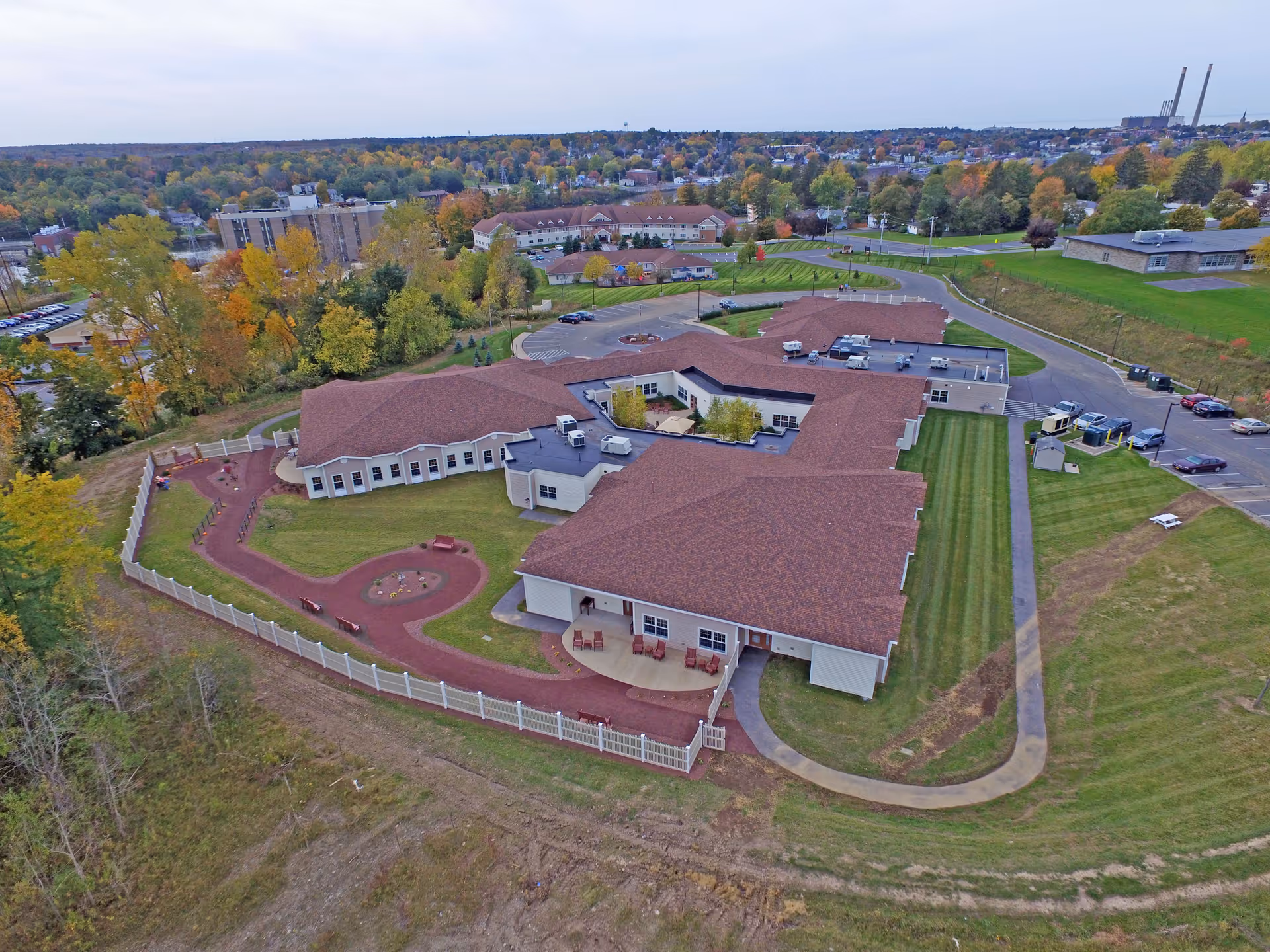 Aerial view of St. Francis Commons Assisted Living Residence showing a large single-story building with a brown roof surrounded by green lawns and a fenced walking path. The surrounding area includes trees with autumn foliage, parking lots, and other buildings in the distance under a cloudy sky.