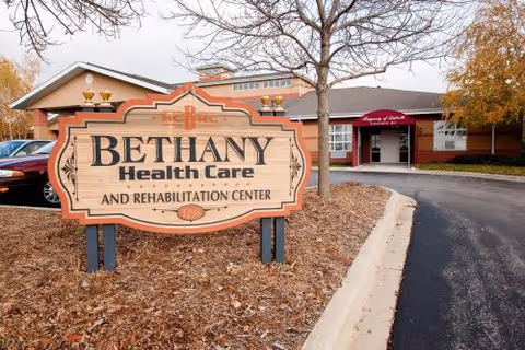 Exterior view of Bethany Health Care and Rehabilitation Center with a large wooden sign in the foreground and the building entrance visible in the background surrounded by trees and parked cars.