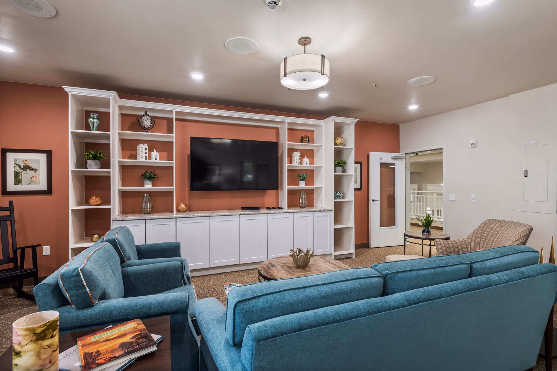 A cozy living room area with teal upholstered sofas and a striped armchair arranged around a wooden coffee table. The back wall features built-in white shelves and cabinets with decorative items and a large flat-screen TV mounted in the center. The walls are painted in a warm terracotta color with white trim, and the ceiling has recessed lighting and a central hanging light fixture. A door with a glass panel leads to another area.
