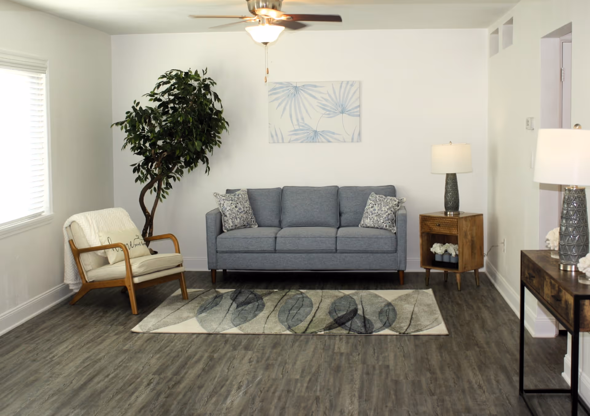 A cozy living room with a gray three-seater sofa adorned with two patterned pillows, a wooden armchair with a white cushion and throw blanket, a leafy green potted plant in the corner, a ceiling fan with a light, a wooden side table with a lamp and decorative flowers, a patterned area rug on wood flooring, and a piece of wall art featuring blue leaf designs.