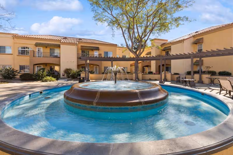 Outdoor courtyard area with a circular water fountain in the center, surrounded by a shallow pool. The courtyard is part of a beige stucco building with two stories, featuring balconies, windows, and a pergola with seating areas. Trees and shrubs are also visible under a partly cloudy sky.