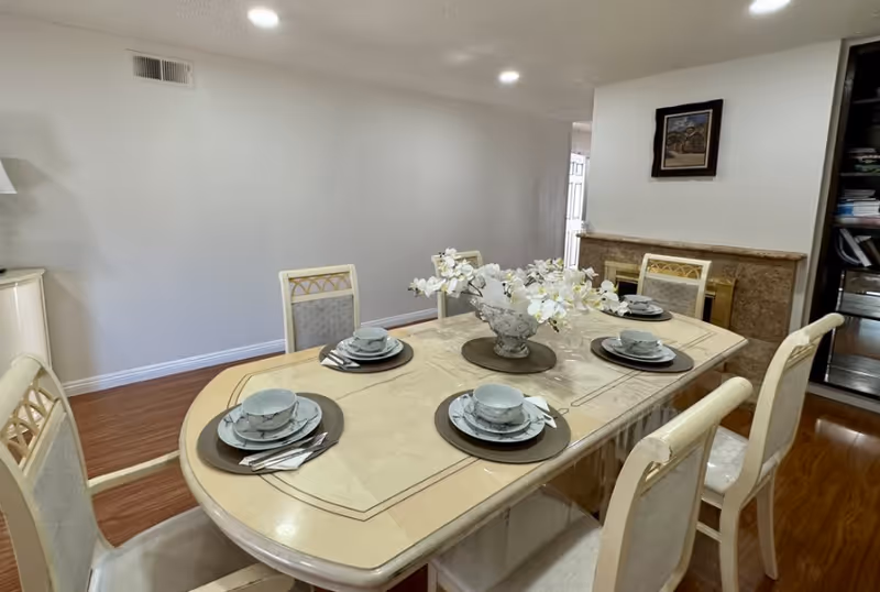 A dining room with a rectangular table set for six people. The table has a beige surface with a glass cover and is decorated with a centerpiece of white flowers in a glass vase. Each place setting includes a plate, bowl, and utensils on a dark placemat. The room has wooden flooring, a fireplace with a framed picture above it, and a bookshelf on the right side.