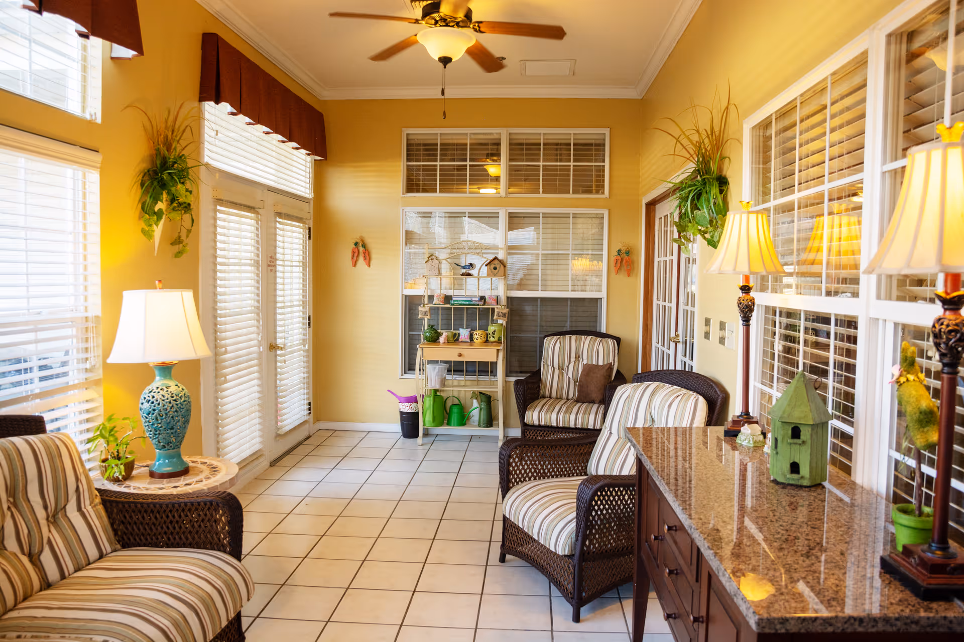 Bright sunroom with wicker armchairs, striped cushions, lamps, potted plants, and a tiled floor.
