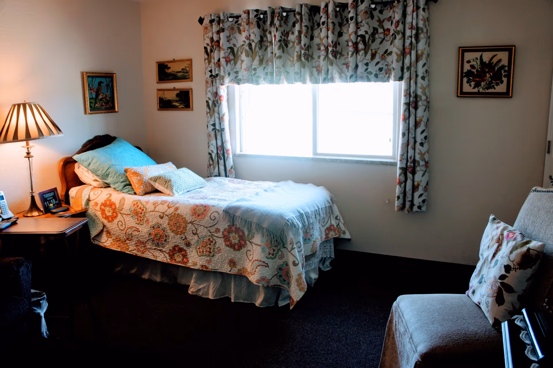 Cozy bedroom with a single bed dressed in a floral quilt, a nightstand and lamp by a large window, and an upholstered chair.