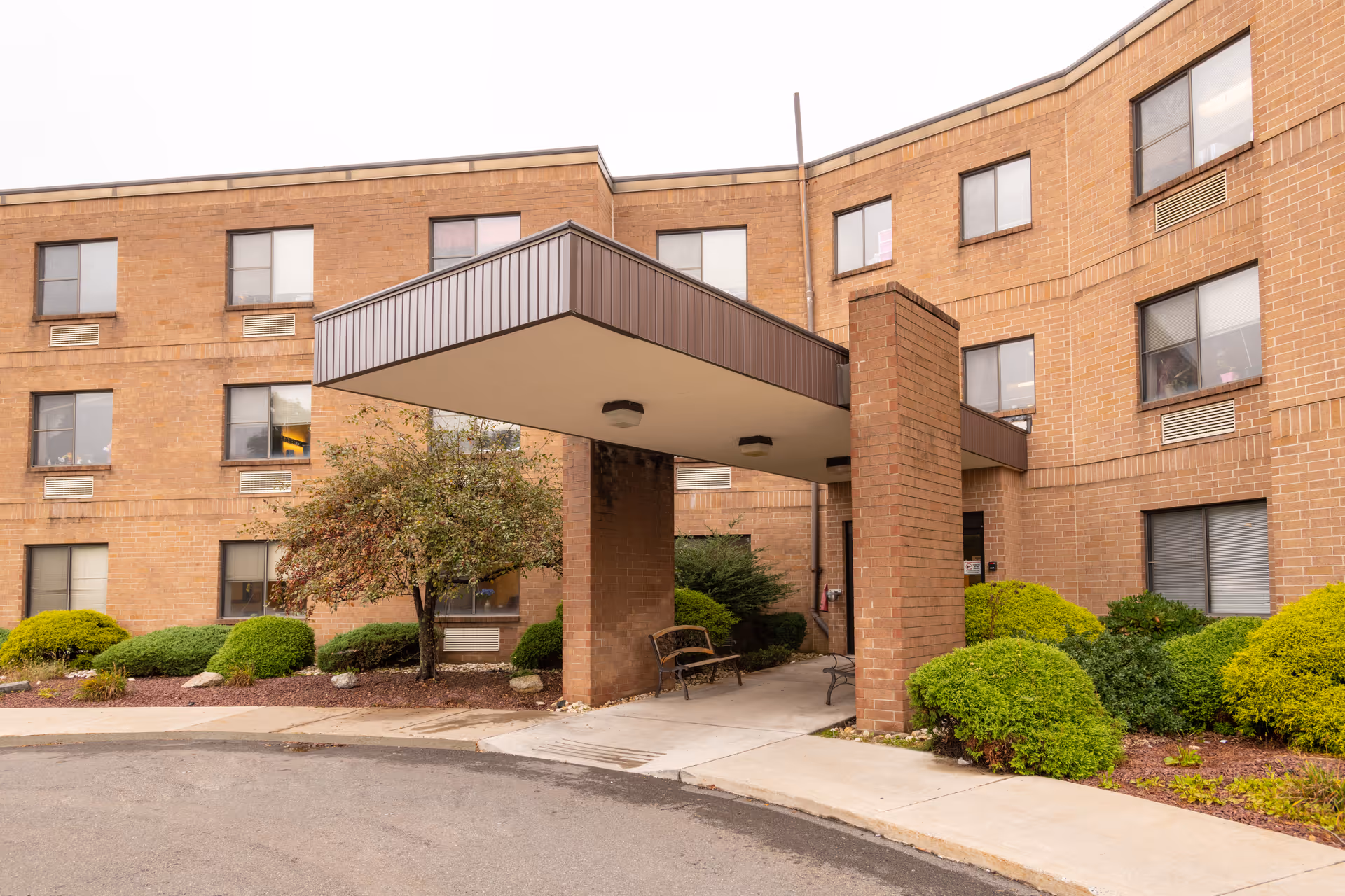 Brick three-story nursing facility entrance with a covered portico, benches, and landscaped shrubs.