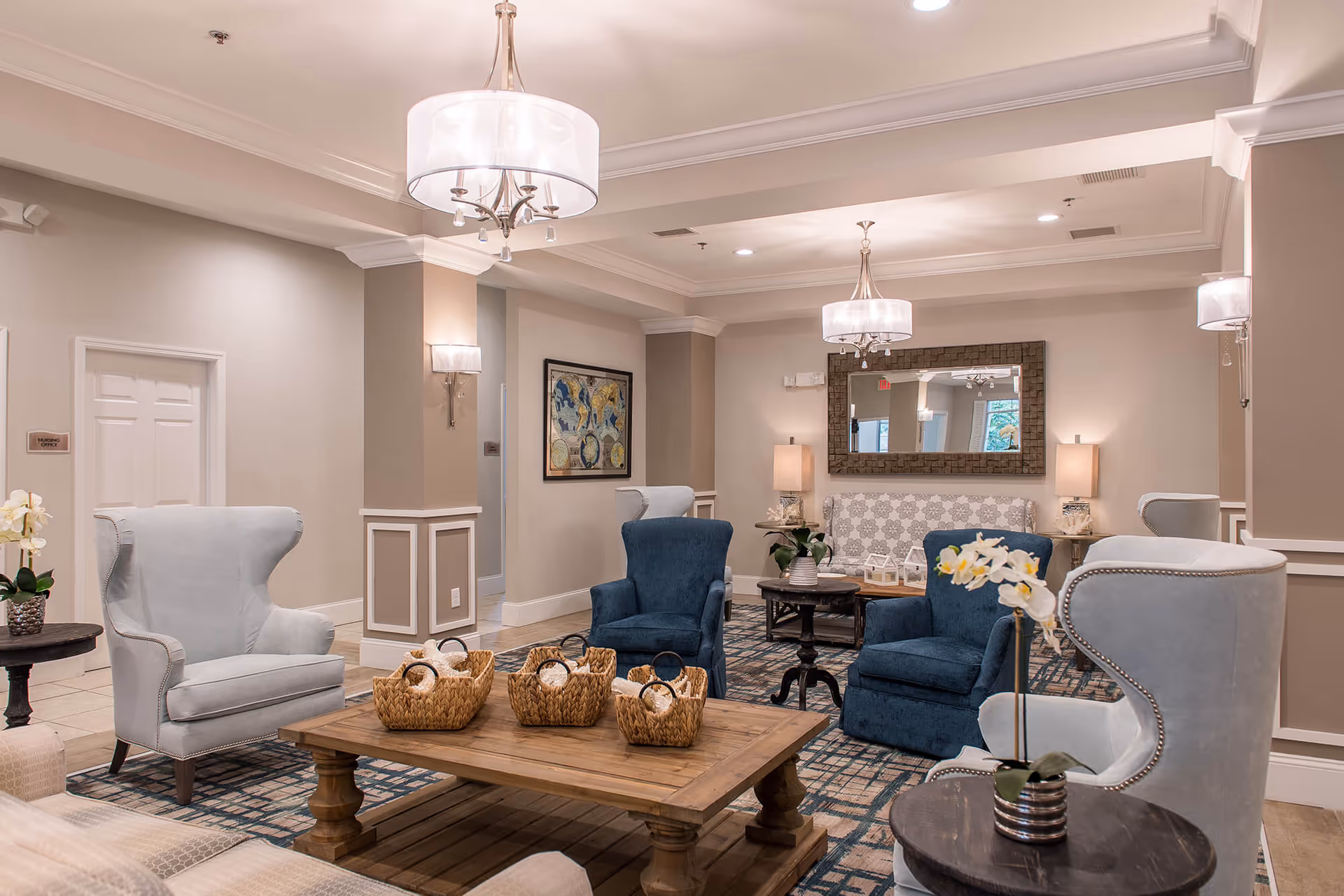 A well-lit, elegant sitting area in a senior living facility with comfortable armchairs in light gray and dark blue, a wooden coffee table with woven baskets, side tables with potted orchids, a patterned rug, wall sconces, a large mirror, and framed artwork on the wall.