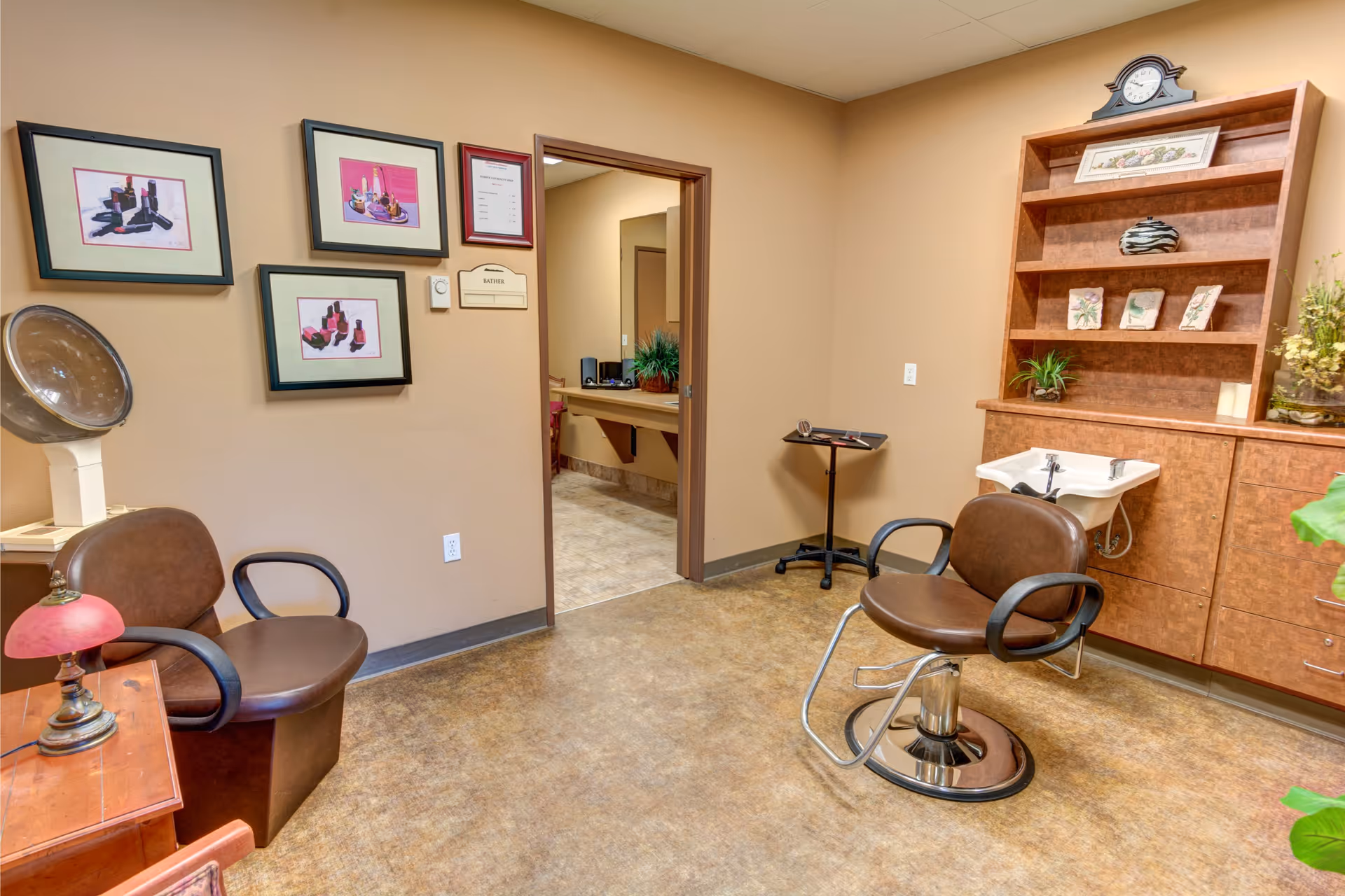 Small salon-style room with two styling chairs, a sink and shelving, framed artwork on the wall, and a doorway to another room.