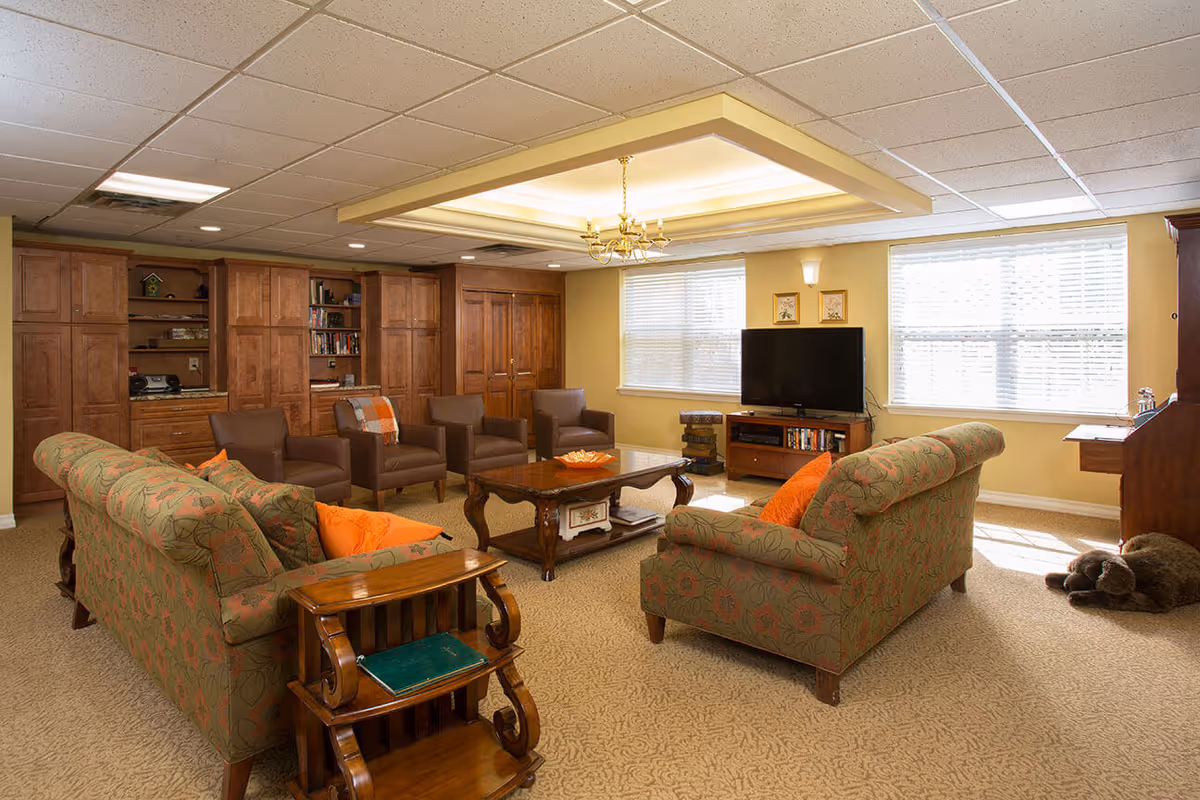 A comfortable communal living room with patterned sofas and armchairs arranged around a coffee table, a television, and wooden built-in cabinets under recessed ceiling lighting.