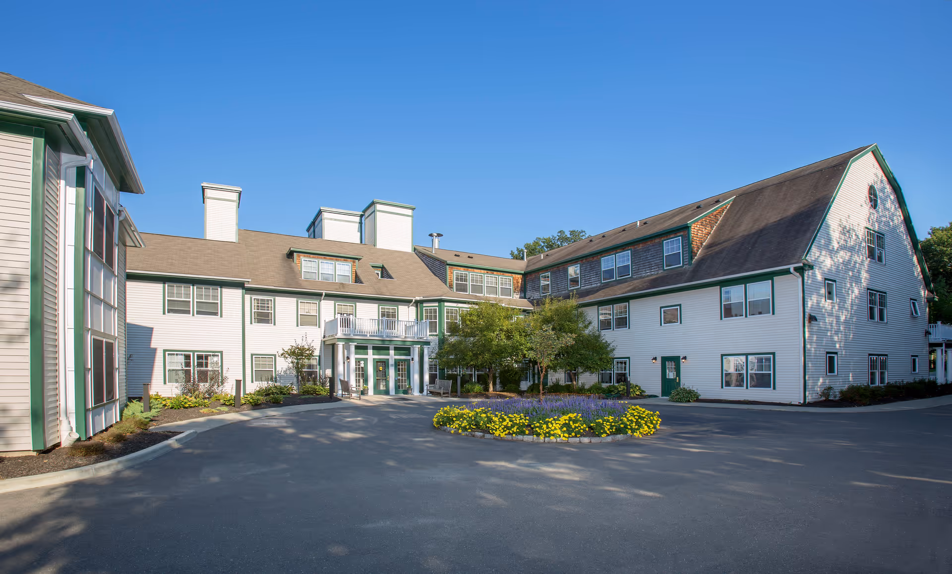 Front exterior of a multi-story white senior living building with green trim, a circular flower bed, and a paved driveway under a clear blue sky.