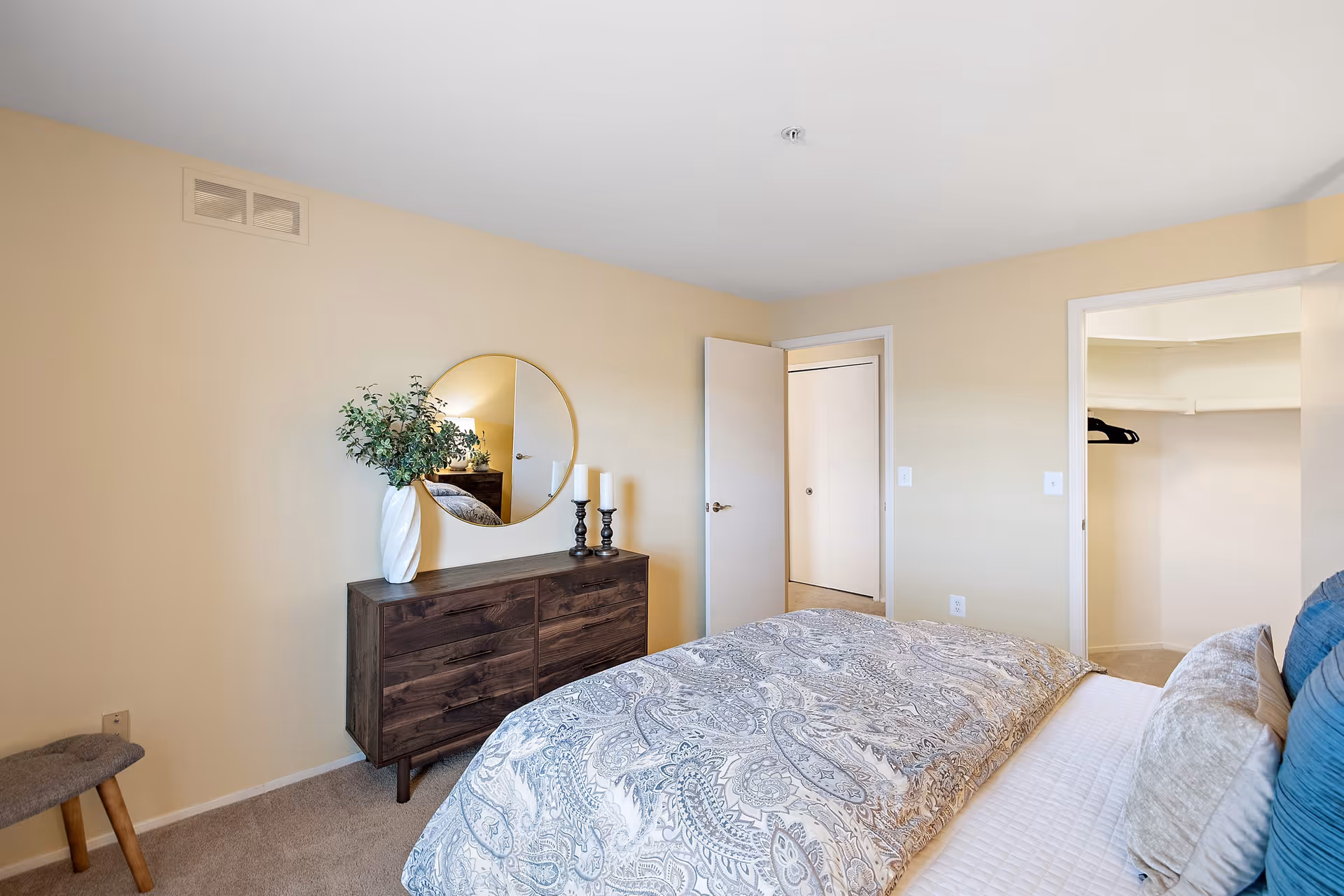 A bedroom with beige walls and carpeted floor featuring a bed with patterned bedding and multiple pillows. There is a dark wooden dresser with a round mirror above it, a white vase with green foliage, and two candle holders. An open door reveals a walk-in closet with shelves and hangers.