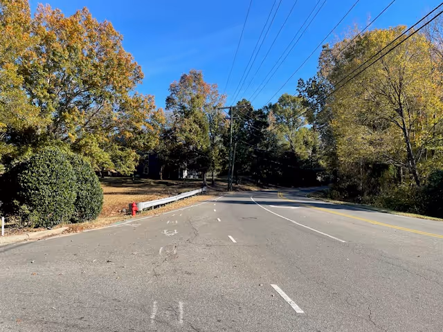 A quiet road lined with trees showing autumn foliage under a clear blue sky. There are power lines running along the right side of the road and a red fire hydrant on the left near a guardrail.