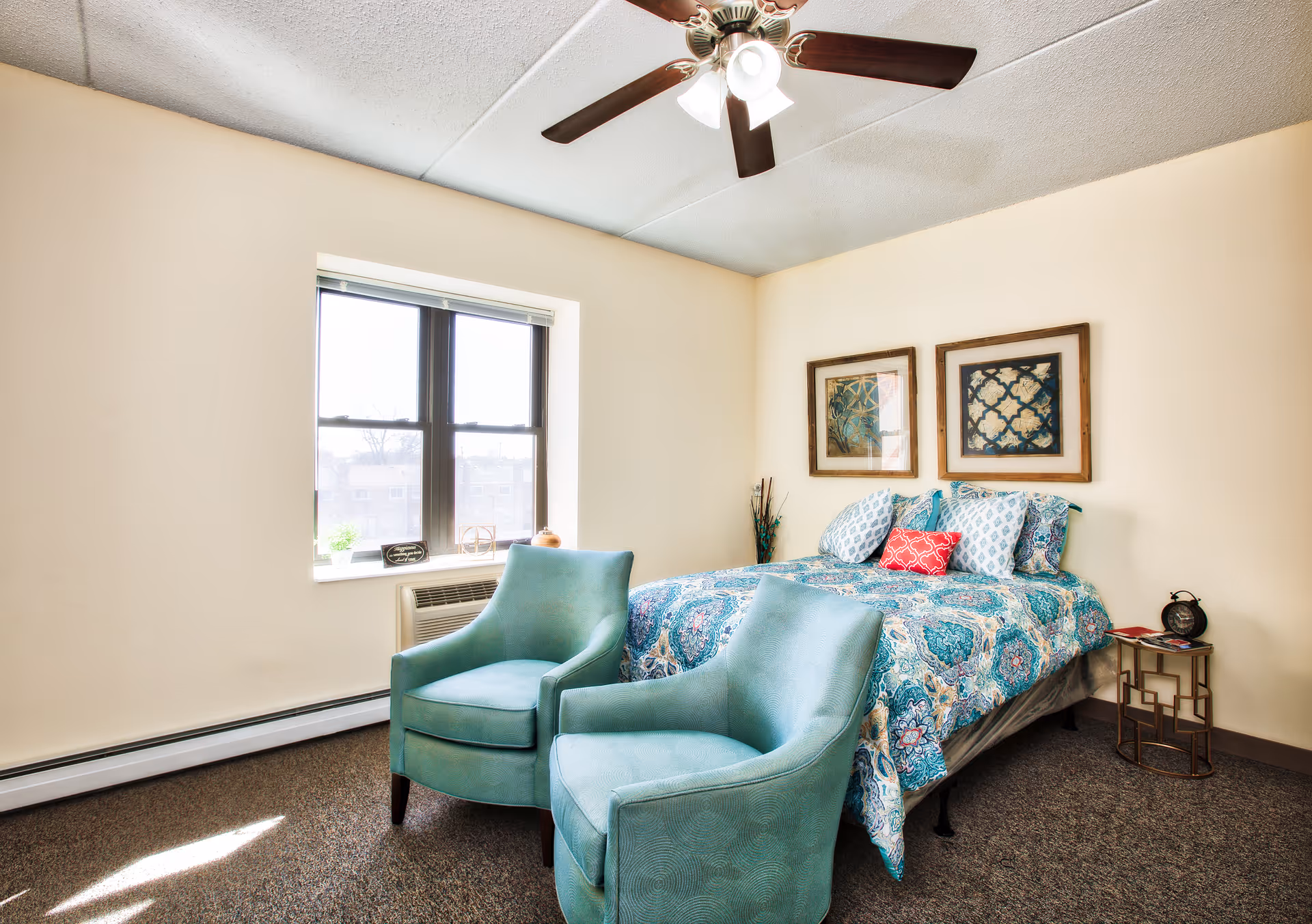 A furnished bedroom with a patterned bed, two teal armchairs, a window, and a ceiling fan.