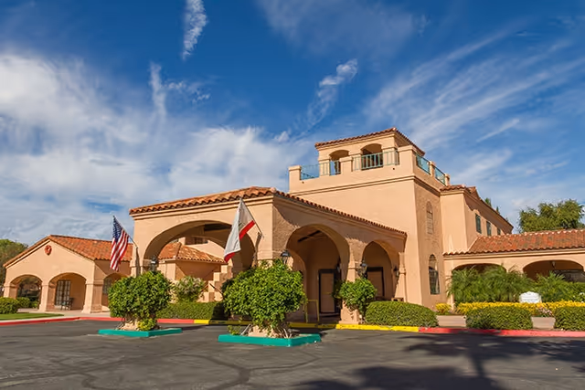 Front entrance of a Spanish-style senior living building with arched entries, flags, and landscaped driveway under a blue sky.