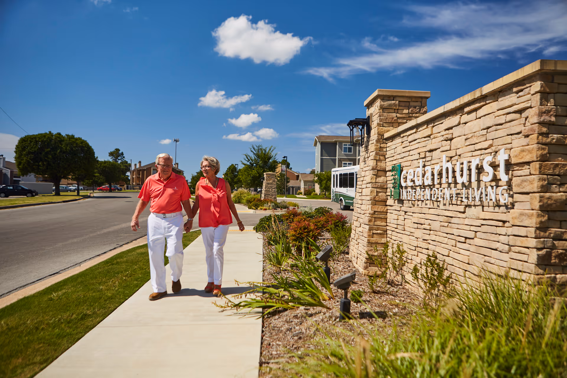 An elderly couple holding hands and walking on a sidewalk next to a stone wall with the sign 'cedarhurst INDEPENDENT LIVING' under a bright blue sky with scattered clouds.