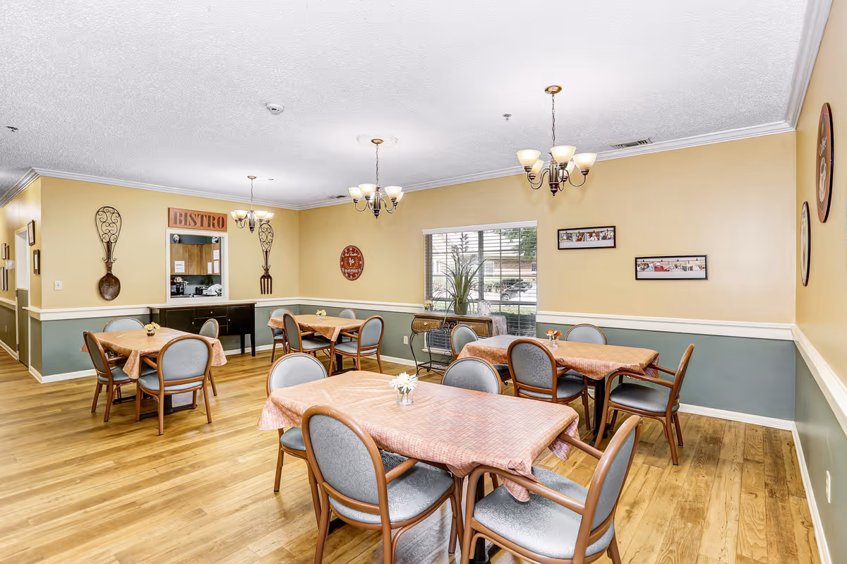 A dining room with several tables covered in pink tablecloths, each surrounded by four chairs with gray cushions. The room has light yellow walls with a green lower half and wooden flooring. There are chandeliers hanging from the ceiling, decorative wall art including large spoon and fork sculptures, and a window letting in natural light. A sign above a pass-through window reads 'BISTRO'.