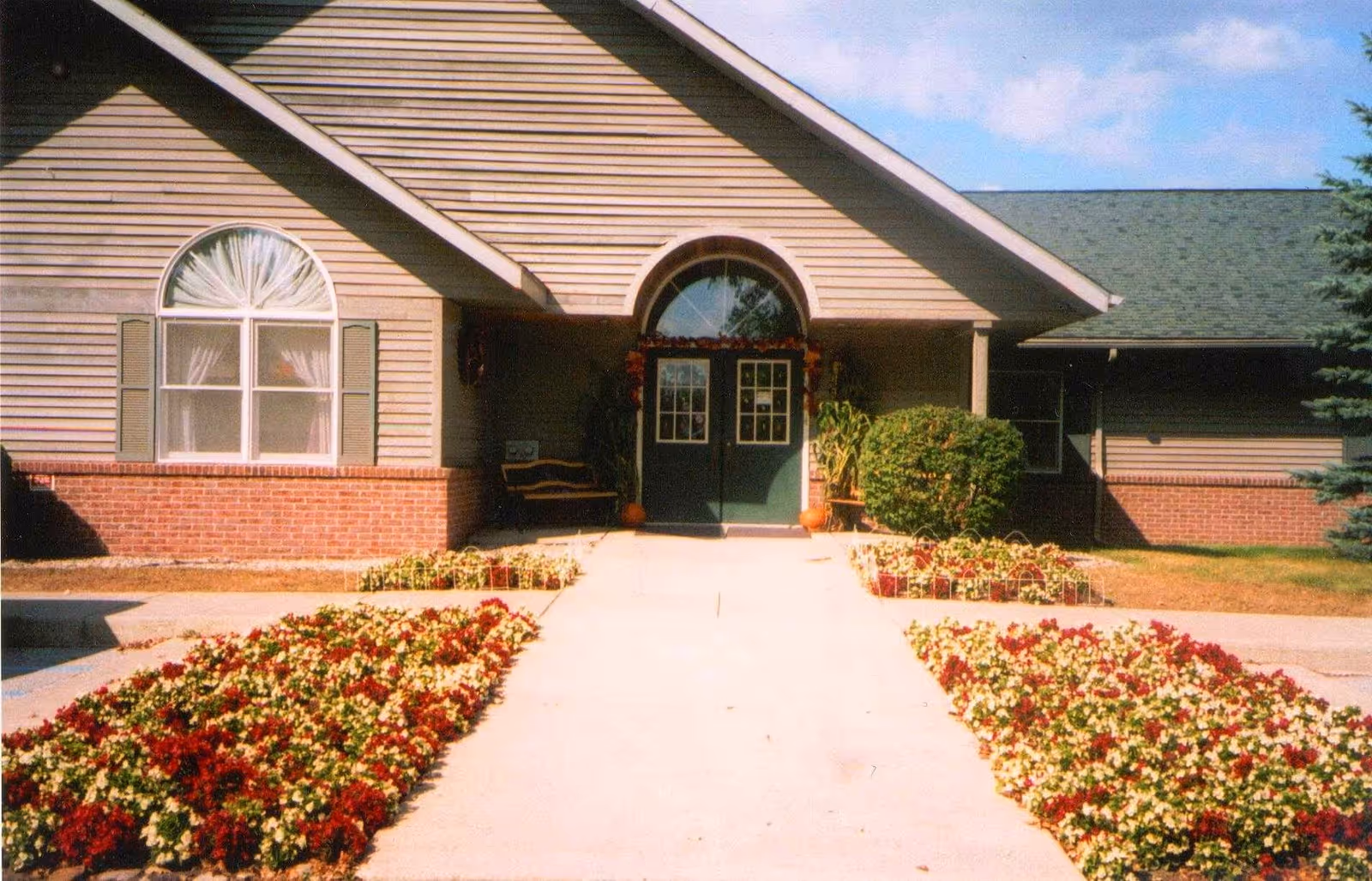 Front entrance of a building with a concrete walkway flanked by flower beds filled with red and white flowers. The building has beige siding with a brick lower half, a large arched window with curtains on the left, and double green doors at the center under a peaked roof. There are bushes and a bench near the entrance.