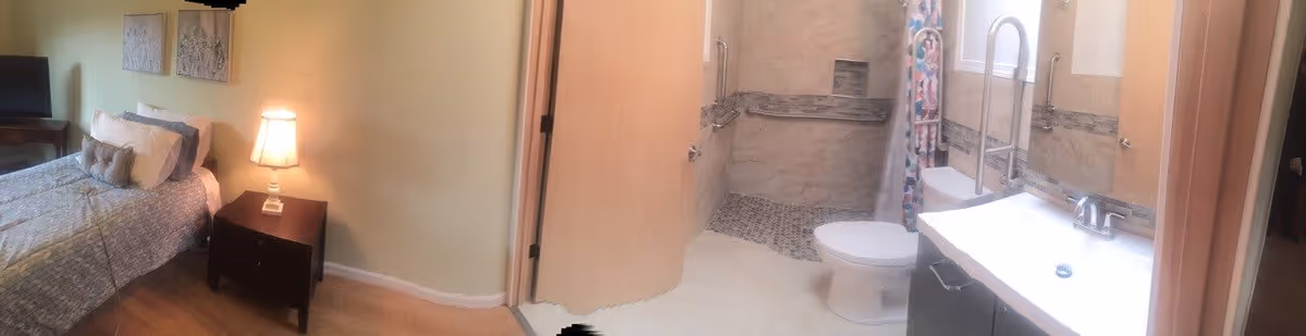 Panoramic view showing a bedroom with a bed and nightstand that opens into an adjacent bathroom with a toilet, sink, and walk-in shower.