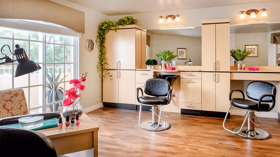 A bright and clean salon area in a senior living facility with two black salon chairs in front of a large mirror and light wood cabinetry. There is a table with a vase of pink flowers, nail polish bottles, and a lamp near a large window with a view of greenery outside. The room has wooden flooring and is decorated with plants and framed pictures on the walls.