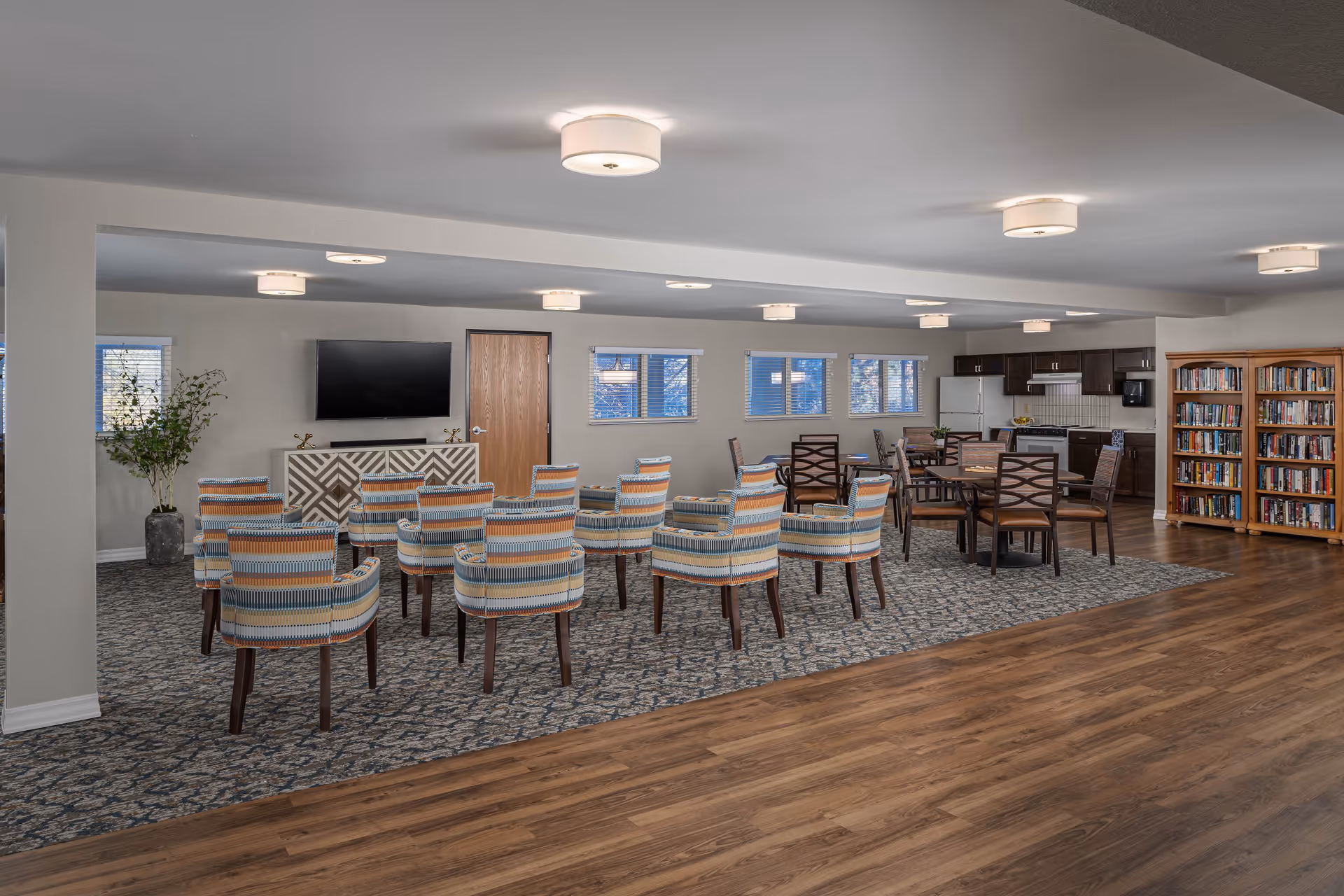 A spacious senior living common area with a seating arrangement of striped upholstered chairs facing a wall-mounted flat screen TV. The room features a patterned carpet section, wooden flooring, a kitchen area with dark cabinets and white appliances, a bookshelf filled with books, and several windows letting in natural light.