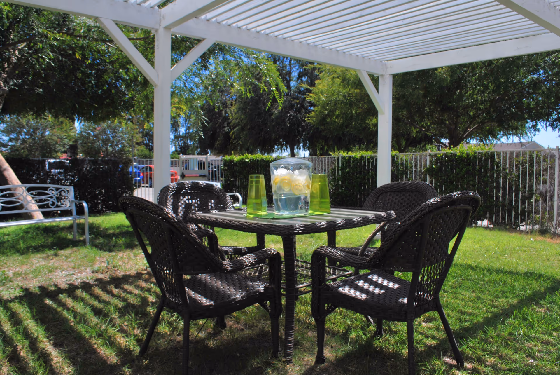 Outdoor seating area with a white pergola overhead, a square table with four dark wicker chairs around it, and a pitcher of lemon water with four green cups on the table. The area is grassy with trees and a white metal bench nearby, enclosed by a white fence.