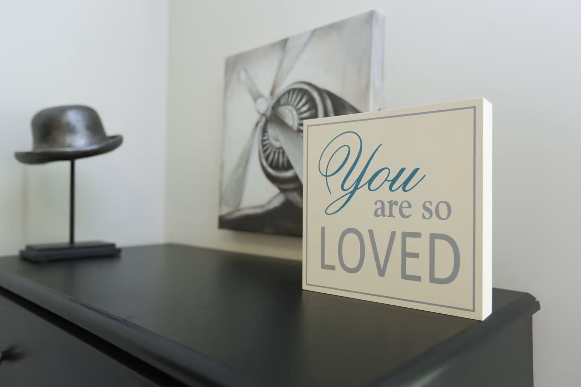 A decorative sign on a black surface with the words 'You are so LOVED' written on it. In the background, there is a black hat on a stand and a black and white painting of an airplane propeller on the wall.