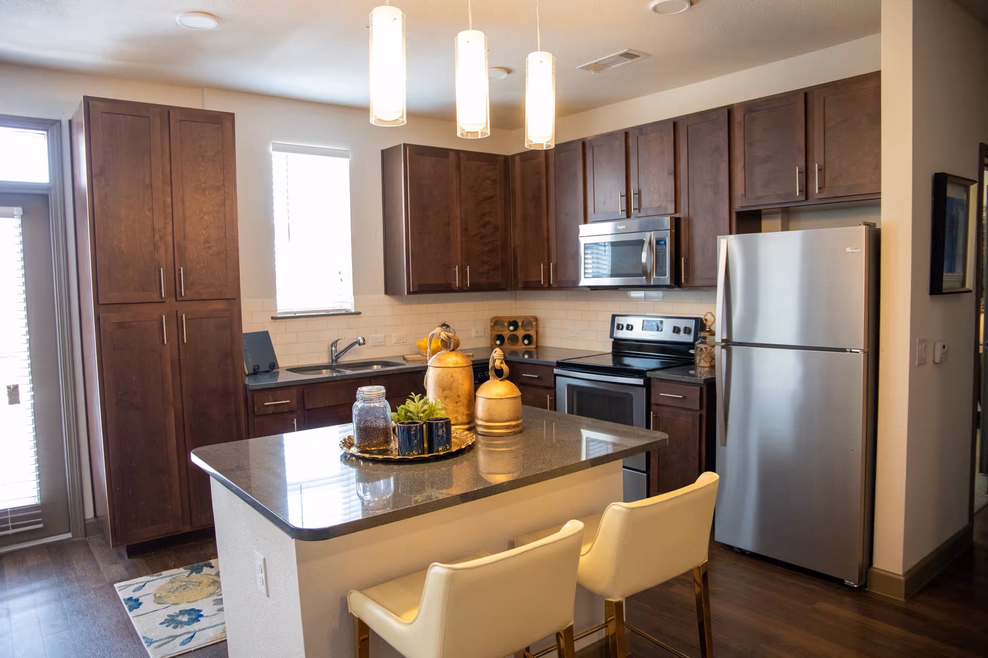 Modern kitchen with dark wood cabinets, stainless steel refrigerator, oven, and microwave. A kitchen island with a dark countertop has decorative items and two white cushioned chairs. There is a window above the sink and pendant lights hanging from the ceiling.