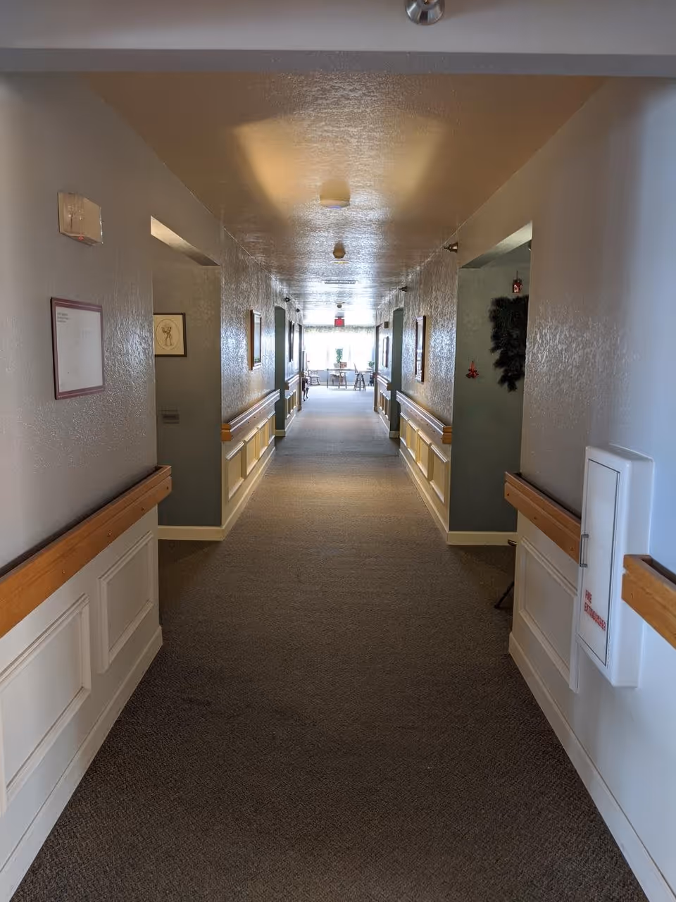 A long, carpeted hallway in a senior living facility with handrails on both sides and framed pictures on the walls. At the end of the hallway, there is a seating area with chairs and a table near a window letting in natural light.