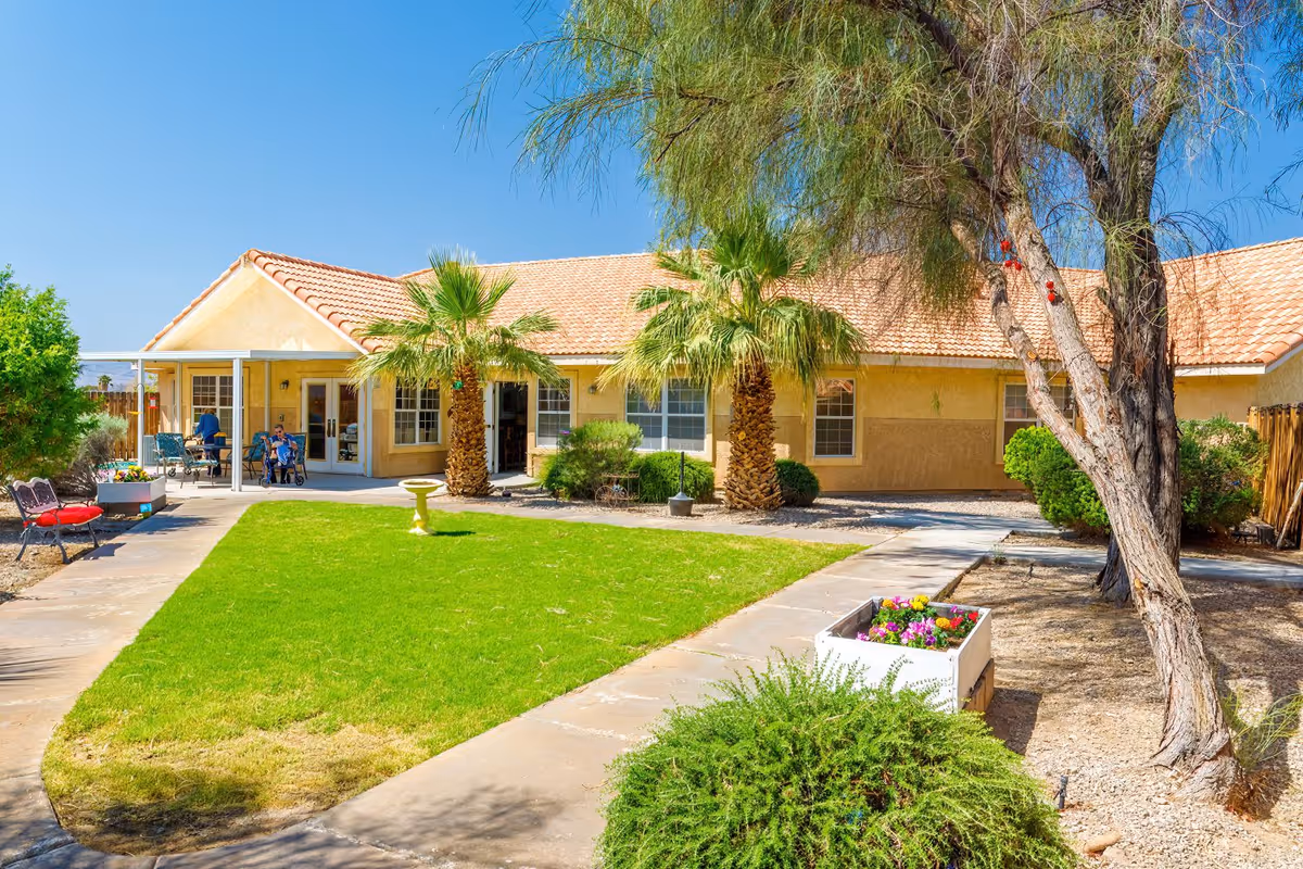 Outdoor courtyard area of Prestige Assisted Living At Lake Havasu featuring a well-maintained green lawn, palm trees, a birdbath, flower planter, benches, and a building with a tiled roof in the background under a clear blue sky.