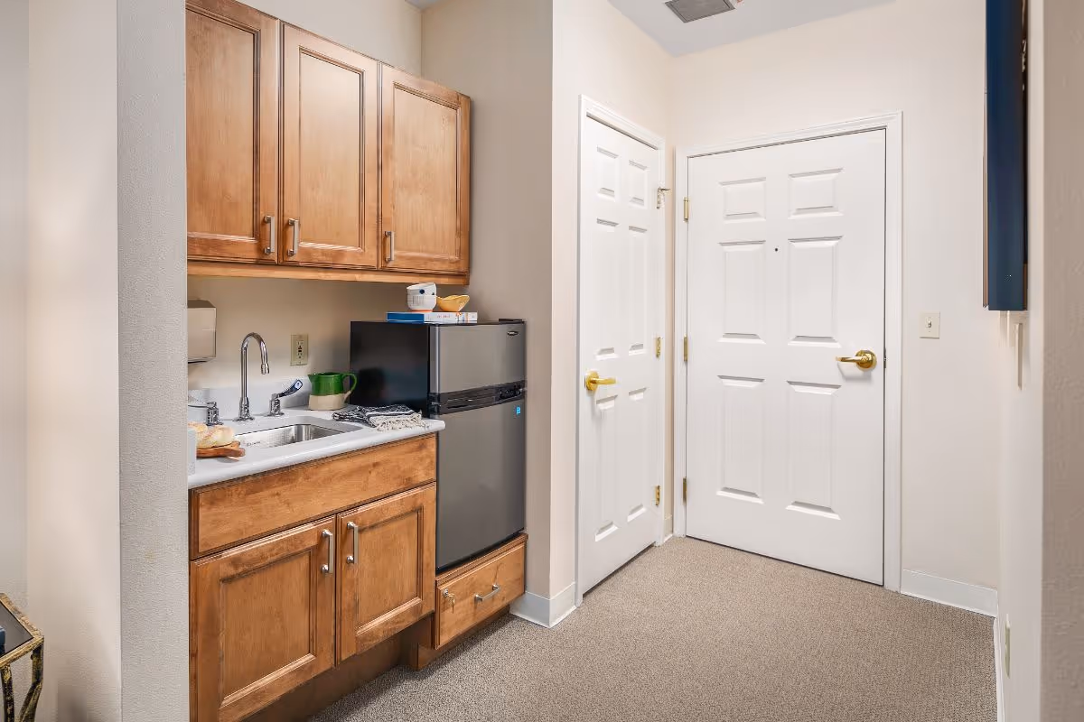 Small kitchenette with wooden cabinets, a sink, mini refrigerator, and two white doors in a carpeted entry area.