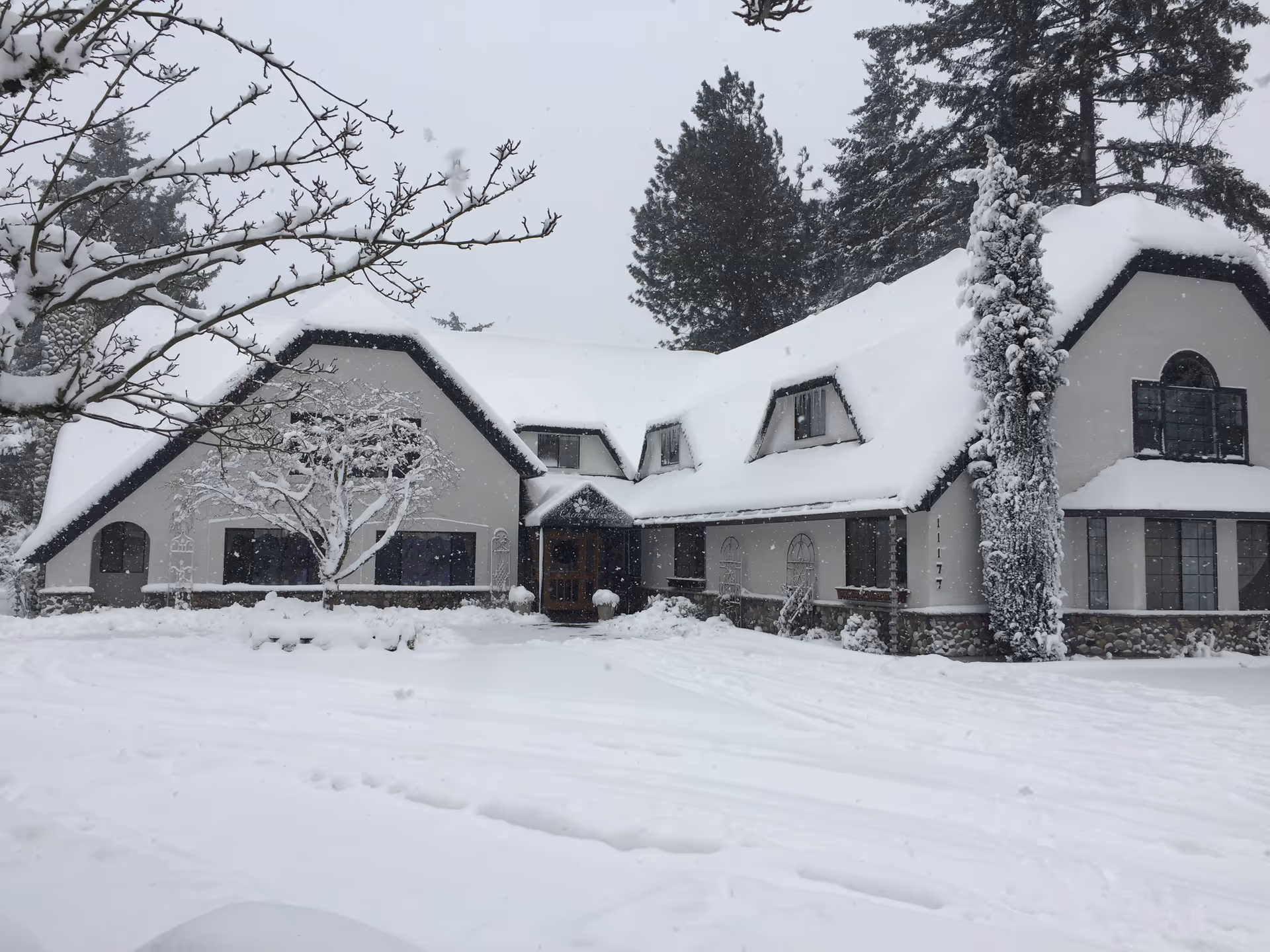 Exterior view of a large residential care facility building covered in snow with snow-covered trees and ground, showing a gray and white house with multiple windows and a steep roof.