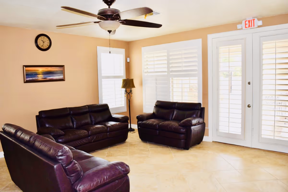 A living room with three dark brown leather sofas arranged around the room. The walls are painted beige, and there is a ceiling fan with lights above. A floor lamp stands near two windows with white plantation shutters. A clock and a framed painting hang on the wall. There is a door with glass panels and an exit sign above it.