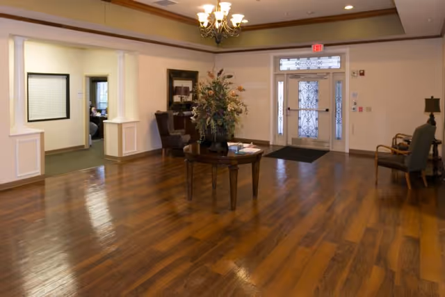 A spacious reception area with polished wooden floors, a central round table with a large floral arrangement, several chairs along the walls, and a glass front door with decorative metalwork.