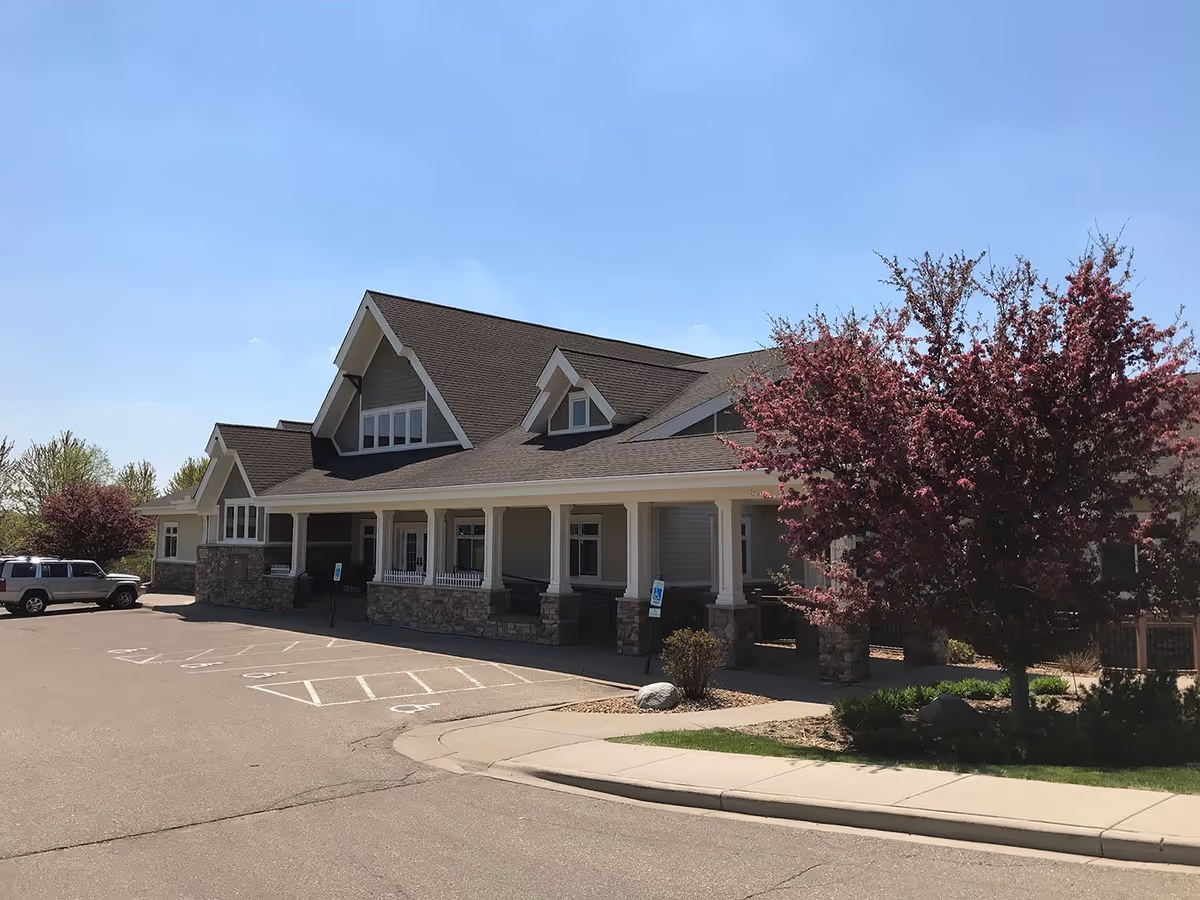 Exterior view of a single-story building with a large covered porch supported by white columns and stone bases. The building has a steeply pitched roof with multiple gables and several windows. There are flowering trees with pink blossoms and a parking lot with marked handicap parking spaces in front of the building under a clear blue sky.