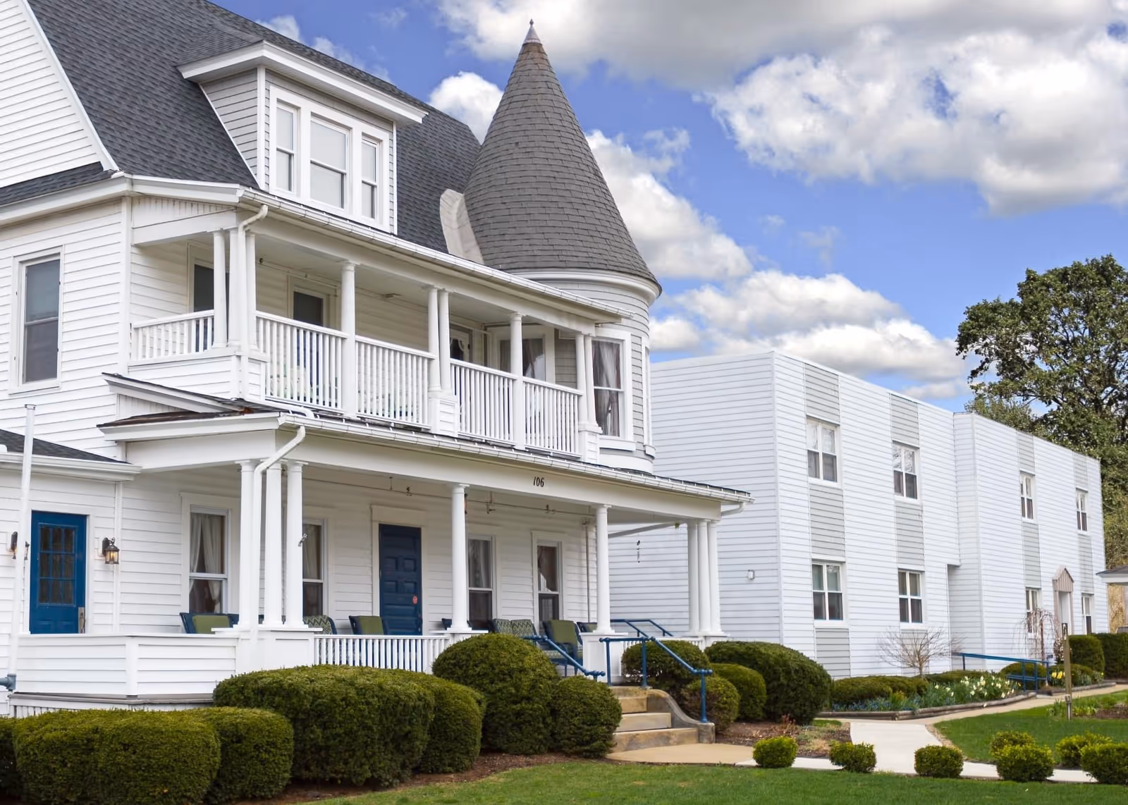 Exterior view of a white senior living facility building with a porch, blue doors, and a turret roof. The building is surrounded by neatly trimmed bushes and a well-maintained lawn under a partly cloudy sky.