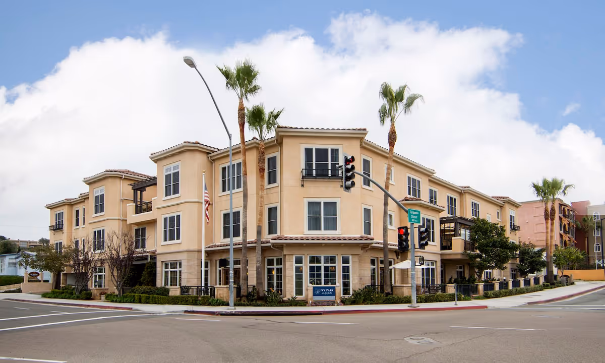Exterior view of a three-story beige building with multiple windows and balconies, palm trees in front, a traffic light, and a street intersection. The building has a sign that reads Ivy Park at La Jolla.