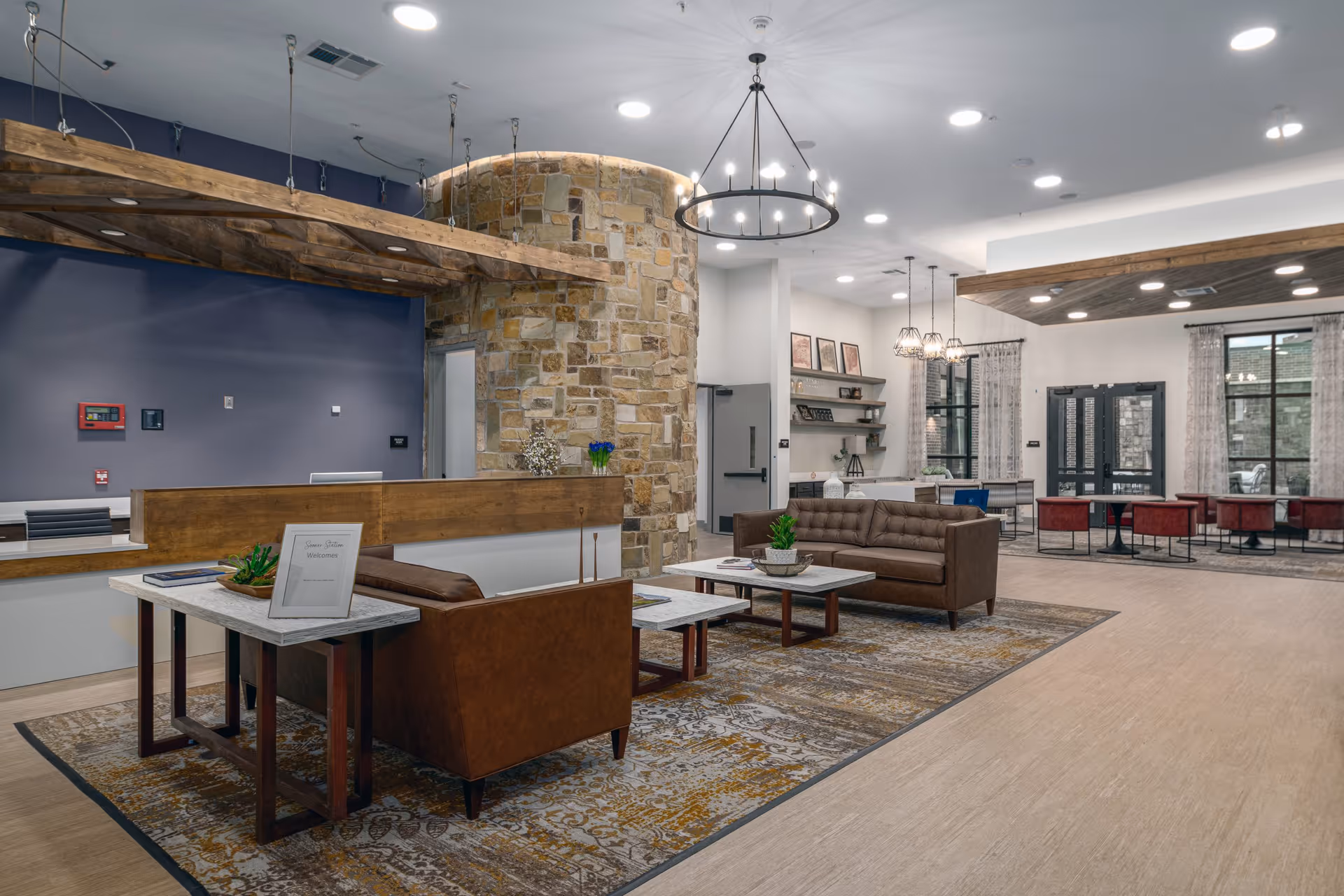 A spacious senior living facility common area with modern decor featuring a stone accent wall, wooden beams on the ceiling, and a circular chandelier. The room includes brown leather sofas, marble-top coffee tables, a patterned area rug, and a reception desk. In the background, there are large windows with curtains, a dining area with red chairs and tables, and shelves with decorative items.