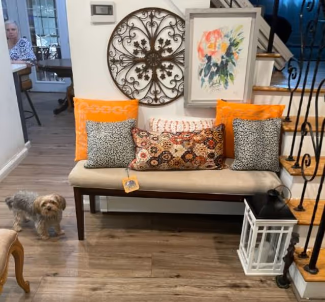 Upholstered bench with colorful decorative pillows and wall art by a staircase, a lantern on the floor, and a small dog on the hardwood floor.