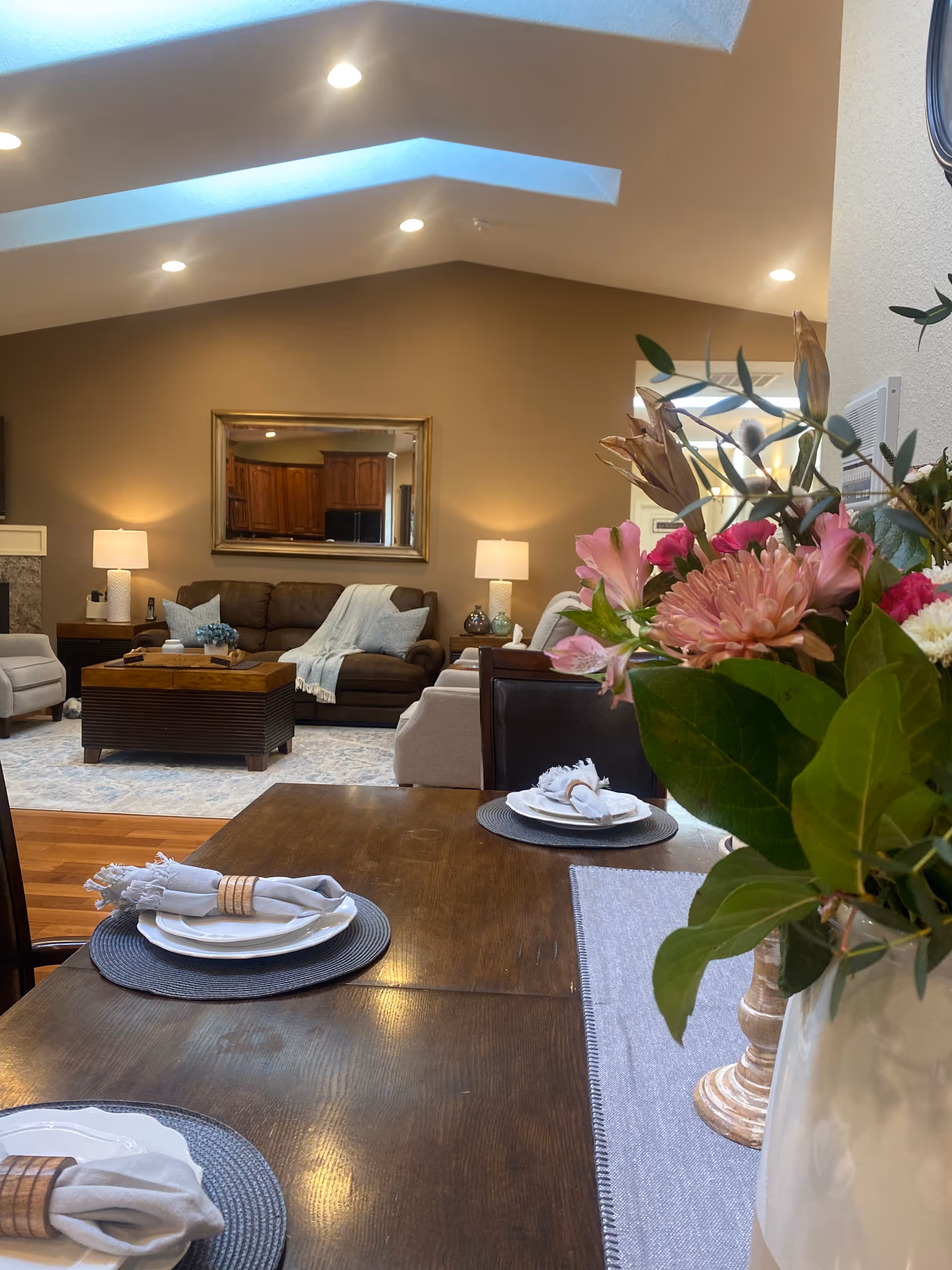 A cozy living room and dining area with a wooden dining table set with plates, napkins, and napkin rings in the foreground. A vase with pink and white flowers is on the table. In the background, there is a brown sofa with a light blue throw blanket, two beige armchairs, a wooden coffee table, two table lamps, and a large mirror on the wall reflecting a kitchen area.
