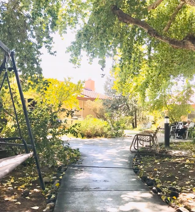 A shaded outdoor pathway in a senior living facility garden area with trees and bushes on both sides. There is a swing on the left and a bench on the right. In the background, a building with a red-tiled roof is visible, along with a person sitting on a chair near the right edge of the image.