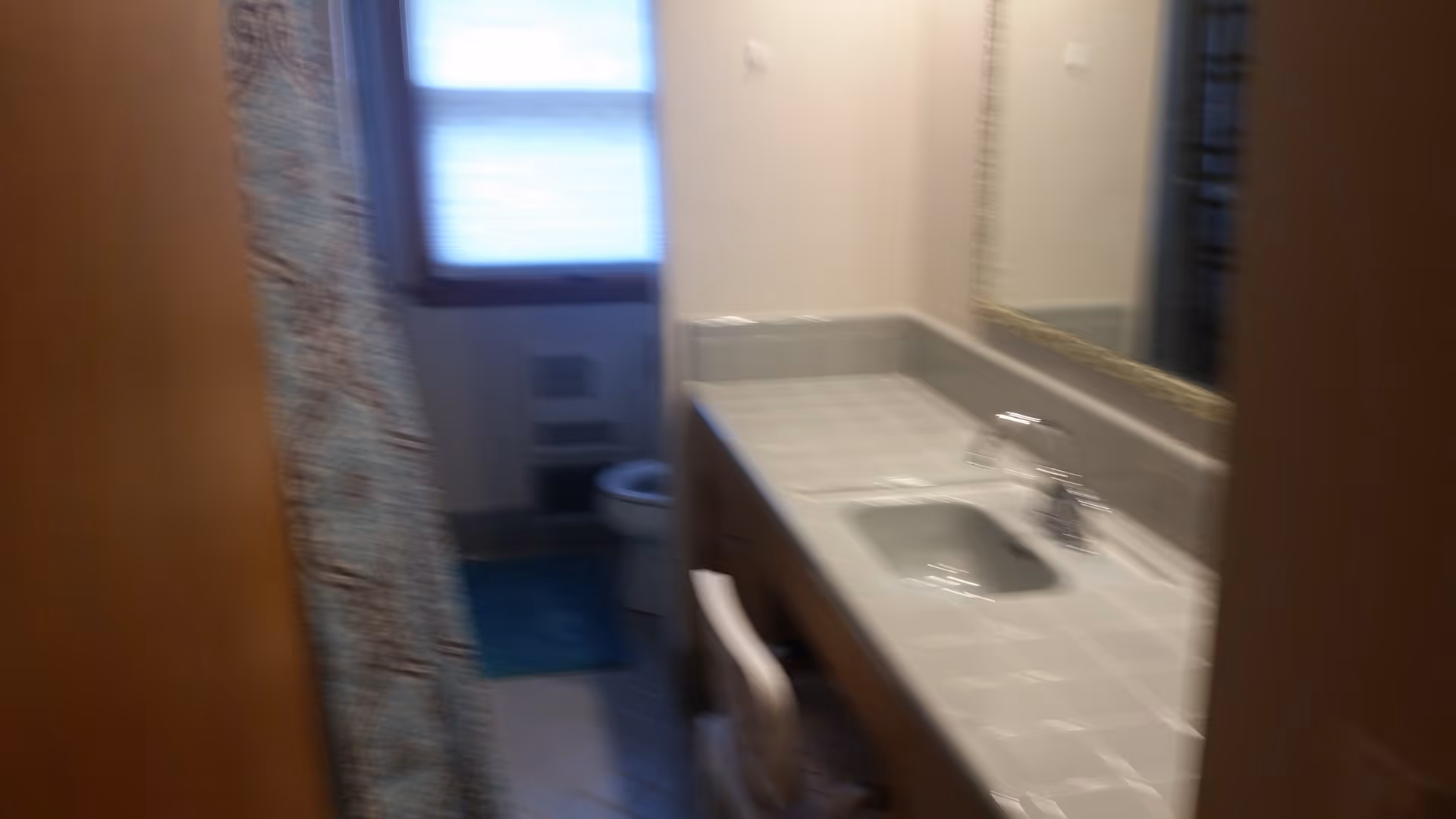 View into a bathroom showing a tiled countertop with a sink and faucet on the right side, a large mirror above the sink, a window with blinds in the background, a toilet, and a shower curtain on the left side.