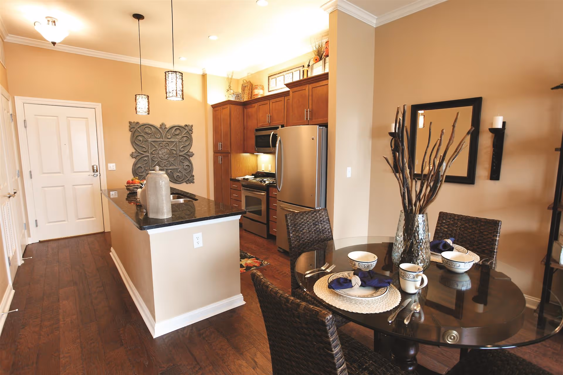 Interior view of a modern kitchen and dining area in a retirement community. The kitchen features wooden cabinets, stainless steel appliances including a refrigerator and stove, and a black granite countertop island with decorative items. Adjacent to the kitchen is a round glass dining table set with four wicker chairs, place settings with plates, bowls, mugs, and blue napkins, and a vase with decorative branches. The walls are painted beige, and there are pendant lights hanging above the kitchen island.