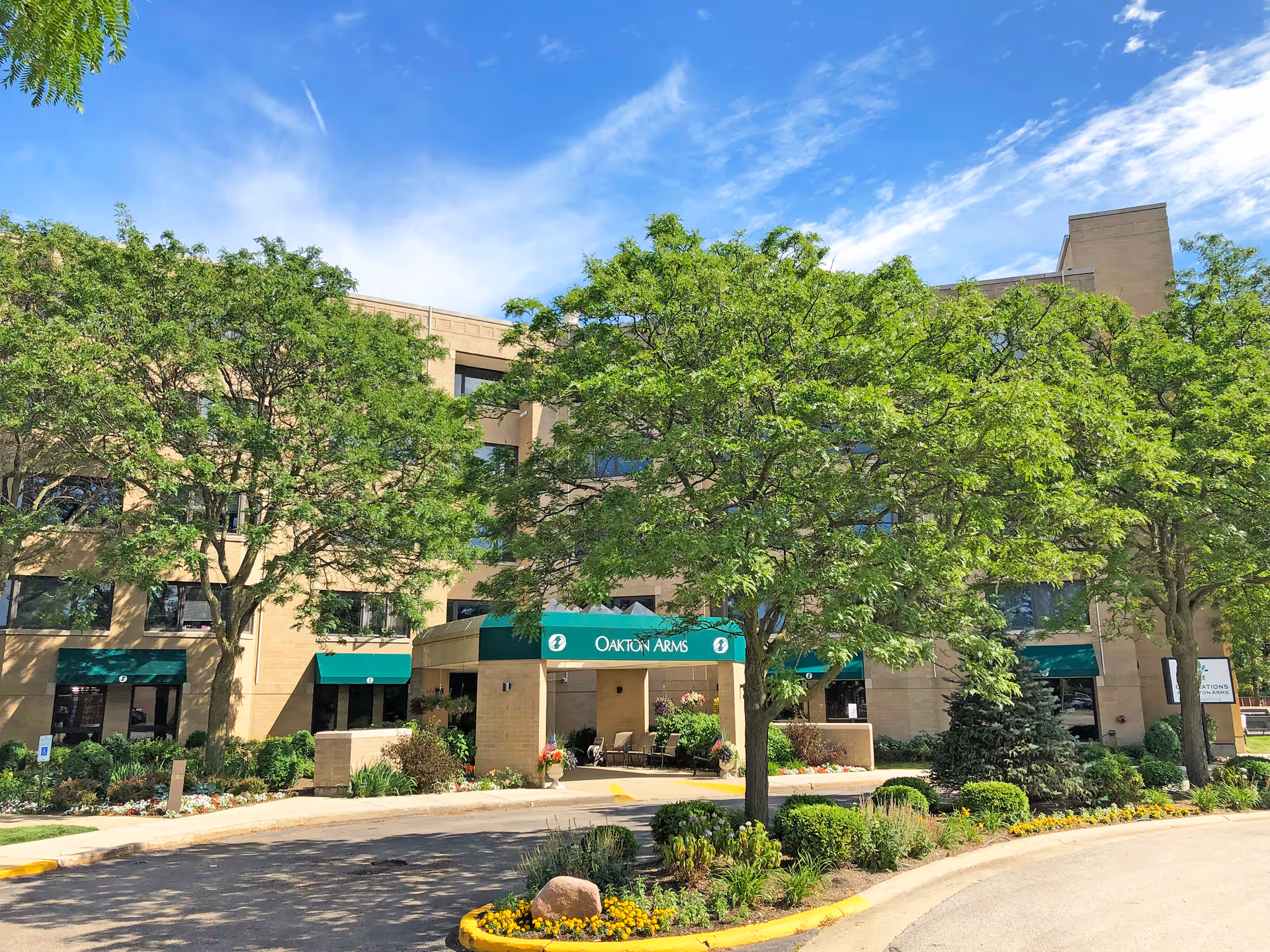 Exterior view of a multi-story senior living facility named Oakton Arms with green awnings over the windows and entrance. The building is surrounded by trees, shrubs, and landscaped flower beds under a blue sky with some clouds.
