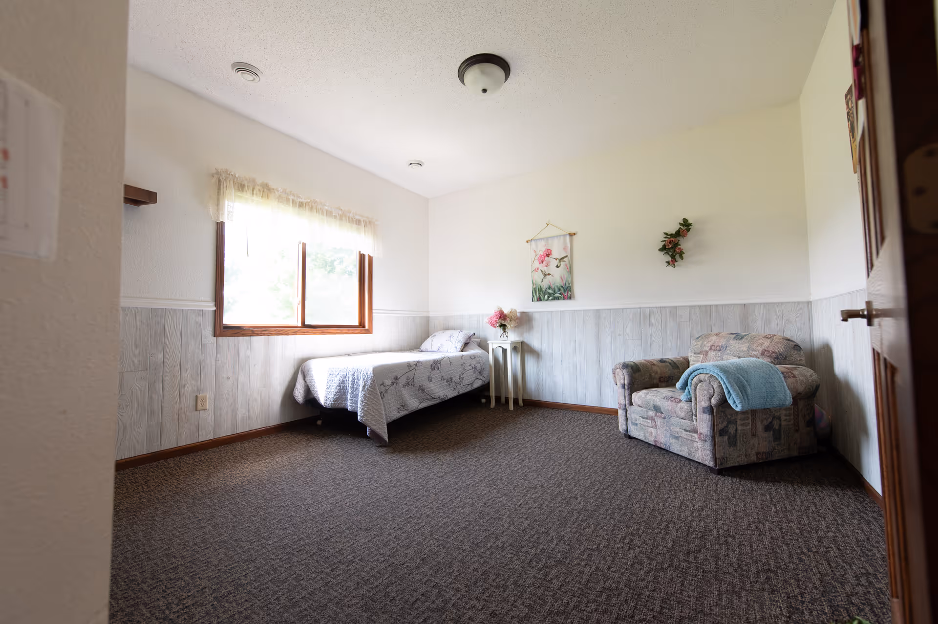 A simple bedroom with a single bed covered in a white and gray patterned bedspread, a small white side table with a flower vase, a floral armchair with a blue blanket draped over it, a window with sheer curtains letting in natural light, and light-colored walls with wood paneling on the lower half.