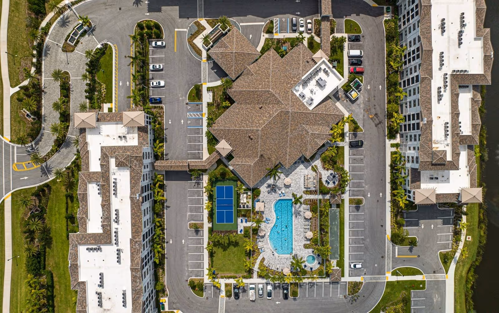 Aerial view of Arcadia Gardens showing a central clubhouse with swimming pool, a tennis court, parking areas, and surrounding apartment buildings.