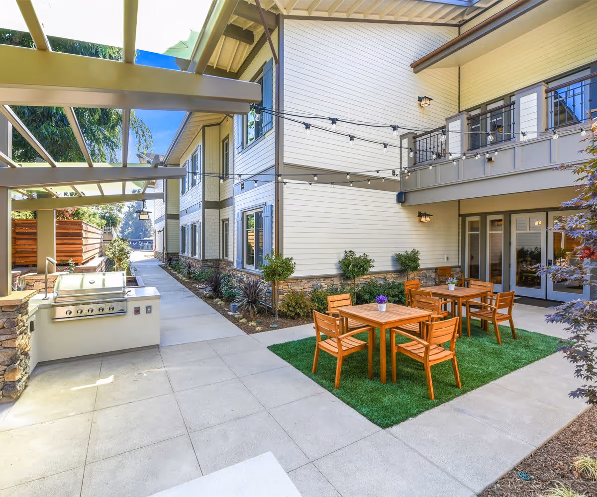 Outdoor courtyard with wooden tables and chairs on a small turf area, a built-in grill under a pergola, string lights, and the facility's exterior walls and balcony.