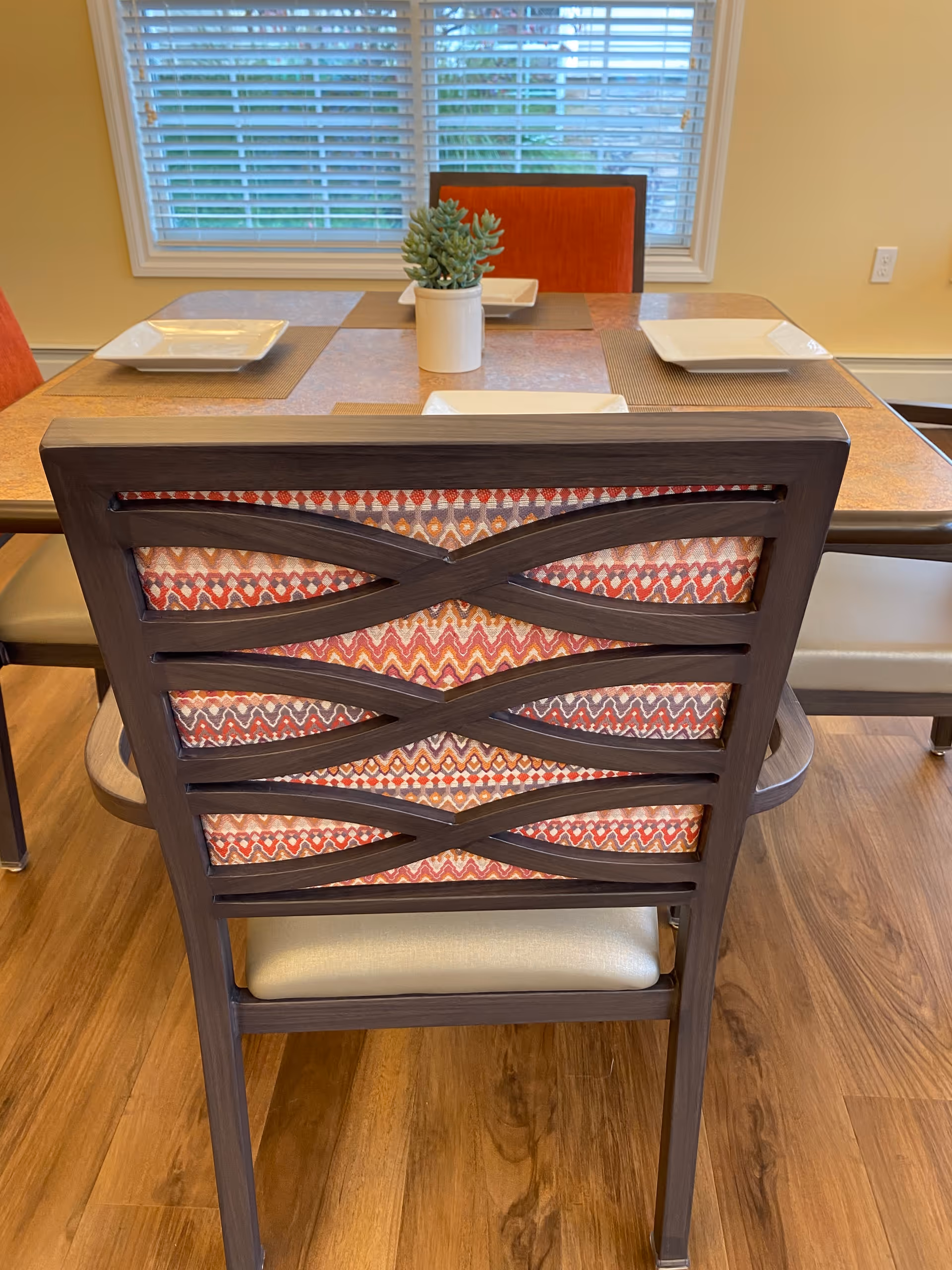 Rear view of a patterned dining chair at a set table with plates and a small potted plant in front of a window with blinds.