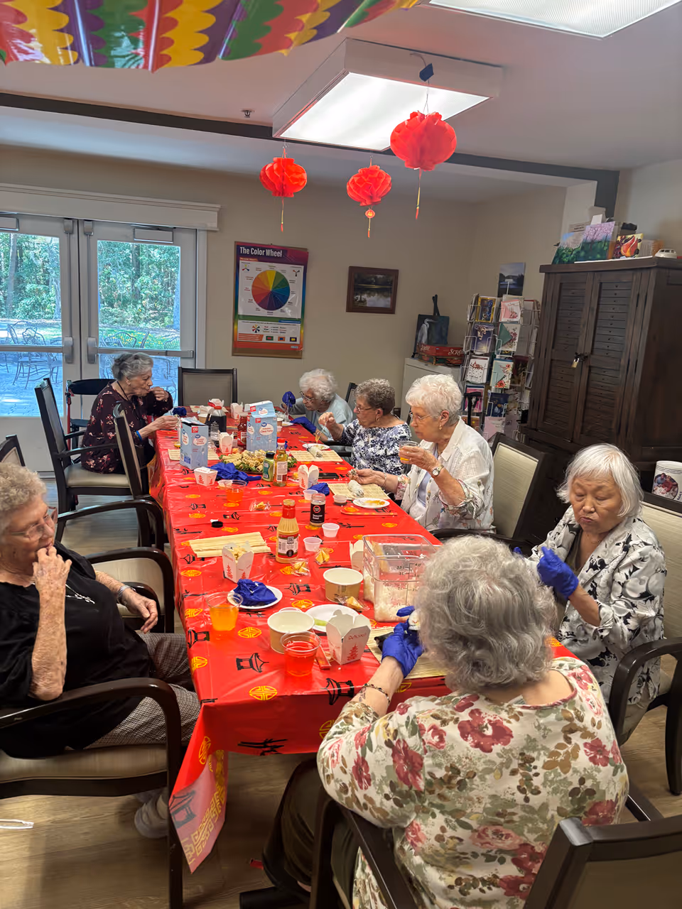A group of elderly women seated around a red-decorated table in a communal dining/activity room eating and crafting.