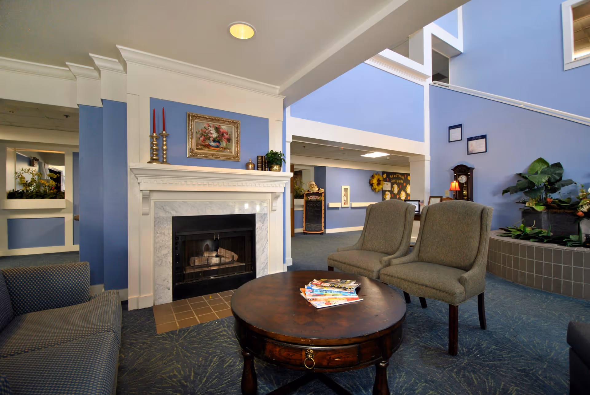 A cozy living room area in Porthaven Manor featuring a white fireplace with a floral painting and two red candles on the mantel. There are two gray upholstered chairs and a round wooden coffee table with magazines on it. The walls are painted blue, and there is a large plant arrangement near a staircase with a grandfather clock and framed pictures on the wall.